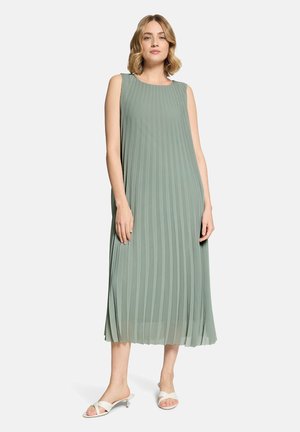 Kobieta w bezrękawowej, zielonkawej sukience midi z plisami, w połączeniu z białymi sandałami na obcasie z odkrytymi palcami, stojąca na jednolitym tle.