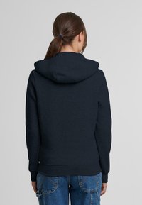 Felpa navy con tasca a marsupio, polsini e orlo a costine. Presenta un cappuccio a doppio strato e una finitura in tessuto morbido e testurizzato.