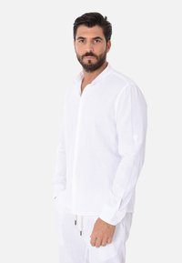 Camicia bianca in lino a maniche lunghe con colletto alla francese, chiusura frontale con bottoni e maniche arrotolate. Esibita su uno sfondo neutro.