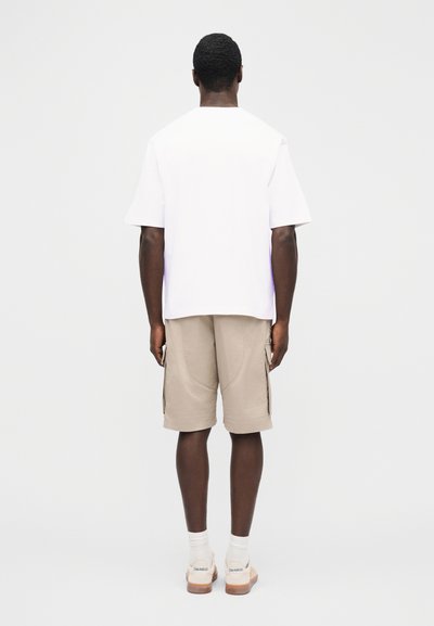 Mann steht mit dem Rücken zur Kamera, trägt ein schlichtes weißes Kurzarmshirt, beige Cargoshorts, weiße Socken und weiße Turnschuhe.