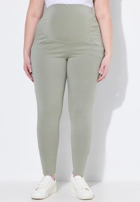 Světle zelené legíny s vysokým pasem, bočními kapsami a hladkou texturou, doplněné o bílé tenisky na jednobarevném pozadí.