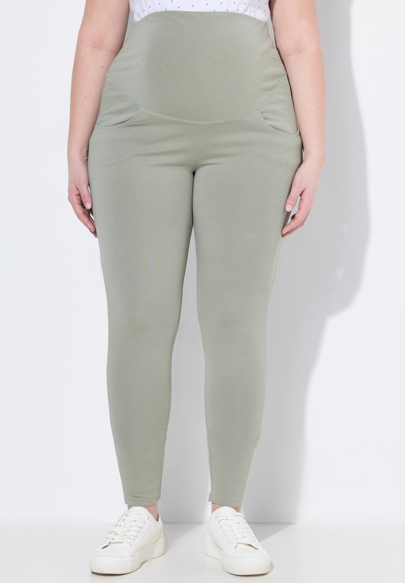 Světle zelené legíny s vysokým pasem, bočními kapsami a hladkou texturou, doplněné o bílé tenisky na jednobarevném pozadí.
