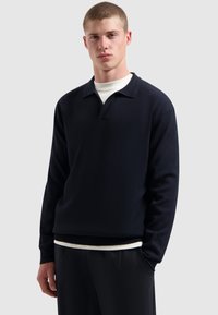 Jonge man met kort haar, gekleed in een donker polo sweater met lange mouwen over een wit shirt en donkere broek, tegen een effen achtergrond.
