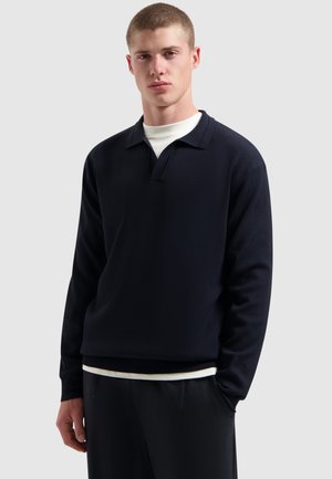 Jonge man met kort haar, gekleed in een donker polo sweater met lange mouwen over een wit shirt en donkere broek, tegen een effen achtergrond.