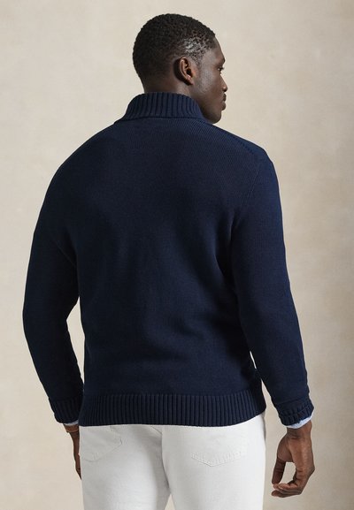 Tamsiai mėlynas megztinis su aukštu apykakliu ir riboto audinio rankogaliais bei apačia. Audinys minkštas, derinamas su baltais kelnėmis.