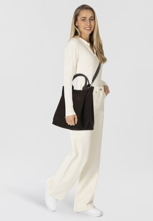 Borsa a tote in pelle marrone scuro con design strutturato e manici sottili. Il modello indossa una maglietta bianca a costine e pantaloni chiari, con sneakers bianche.
