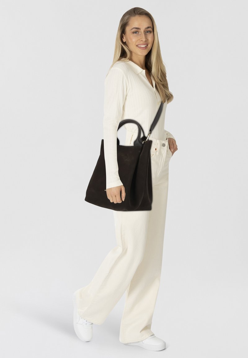 Borsa a tote in pelle marrone scuro con design strutturato e manici sottili. Il modello indossa una maglietta bianca a costine e pantaloni chiari, con sneakers bianche.