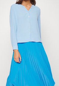Blusa de manga larga azul claro con escote en V y cierre de botones, combinada con una falda plisada de color azul brillante. Textura de tela suave.
