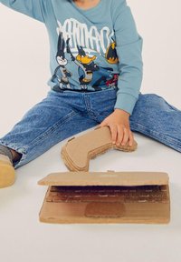 Niño vestido con una sudadera azul clara con personajes de dibujos animados, sentado en el suelo con un portátil de cartón y accesorios de controlador de juego.