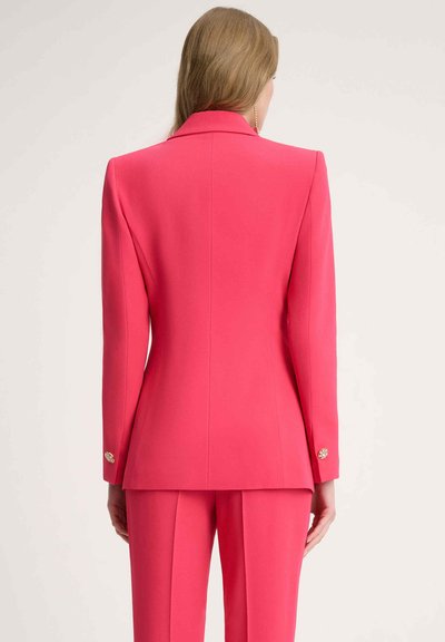 Pink blazer yang fitted dengan kerah notch, bahan halus, dan aksen kancing emas di pergelangan tangan, menampilkan tampak belakang yang disesuaikan.