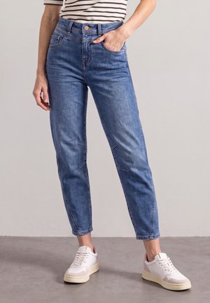 Kvinde iført blå taperede jeans med knæ-sømdetaljer og hvide sneakers, der står på en grå gulv.