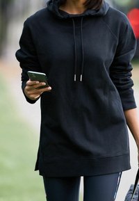 Felpa nera oversize con cappuccio regolabile, con polsini a costine. Chi la indossa tiene uno smartphone e abbina dei leggings aderenti.
