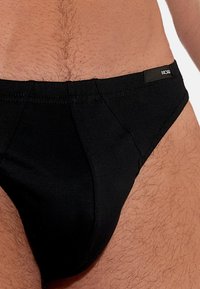 Schwarze Baumwoll-Badebriefs mit niedrigem Schnitt, kontrastierenden Nähten und einem kleinen Logo-Tag am Bund. Weiche Textur mit enger Passform.