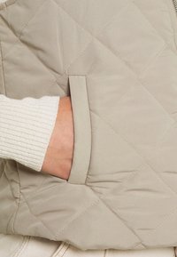 Beżowa pikowana kamizelka z boczną kieszenią. Teksturowany materiał z pionowymi ribbowymi detalami rękawów częściowo widocznymi z lewej strony. Minimalistyczny design.