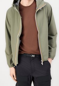 Olivgrön vindjacka med silverdragkedja och orange detalj. Bärs över en brun t-shirt, ihop med svarta cargobyxor.