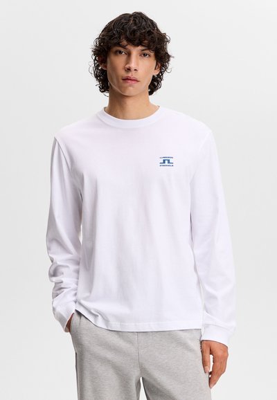 Νέος άνδρας με σγουρά σκουρόχρωμα μαλλιά, φορώντας άσπρο πουκάμισο μακρυμάνικο με μπλε λογότυπο και ανοιχτόχρωμο γκρι παντελόνι, ποζάροντας με το ένα χέρι στην τσέπη.