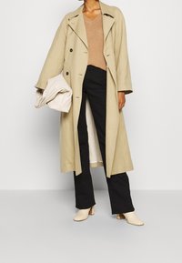 Personne portant un manteau long beige par-dessus un pull marron à col en V et un pantalon noir, tenant un sac beige texturé, debout sur un sol gris.