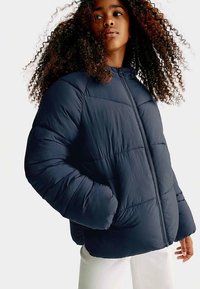 Marineblauwe donsjas met een ritssluiting aan de voorkant, hoge kraag en een rechte pasvorm. Heeft doorgestikte gedeeltes en elastische manchetten voor warmte.