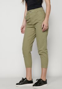 Pantalones de corte tapered en verde oliva con cintura ajustable, bolsillos laterales y una textura suave de algodón. Combinados con zapatillas negras con tachuelas.