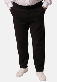 Pantalones negros con una textura suave, diseño ajustado, bolsillos laterales y cierre de botón frontal, combinados con zapatillas blancas.