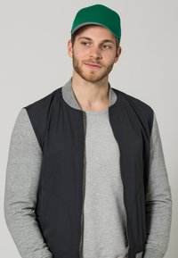 Chaqueta bomber negra con cremallera y mangas grises, llevada sobre una camiseta gris, complementada con una gorra verde con visera gris contrastante.