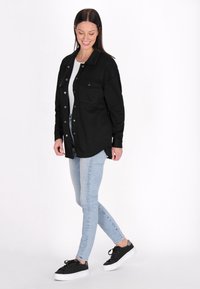 Giacca camicia nera con un motivo strutturato, due tasche sul petto e una vestibilità rilassata, abbinata a jeans tight blu chiaro e sneakers nere.