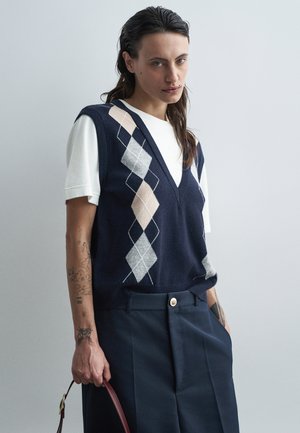Navy sleeveless vest with a diamond pattern in beige and gray, layered over a white t-shirt; paired with high-waisted dark trousers.