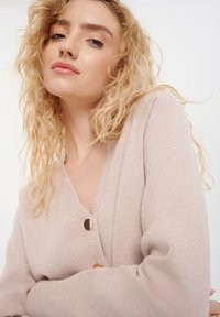 Lichtroze cardigan gemaakt van zachte stof, met een diepe V-hals, lange mouwen en twee opvallende gouden knopen als accenten.