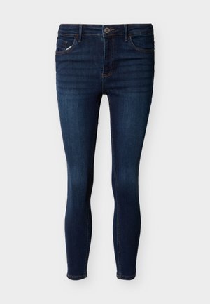 Mørkeblå skinny jeans med knap- og lynlås foran, fem lommer og let falmning på lårene mod en ensfarvet baggrund.