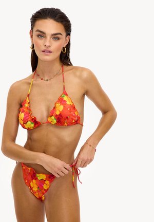 Femme ajustant le lien latéral de son bikini rouge à motif floral avec des fleurs jaunes et orange, portant des boucles d'oreilles anneaux dorés et un collier de perles colorées.