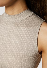 Top senza maniche color beige con colletto alto e un intricato motivo a zigzag in una texture sottile. Tessuto liscio con un design aderente.