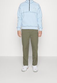 Svetlemodrý svetr s čtvrtzipem a vyšitými detaily, kombinovaný se zelenými olivovými kalhotami a bílými teniskami s červenými detaily.