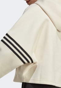 Krótka bluza z kapturem z ecru materiału o fakturowanej powierzchni. Posiada kaptur i czarne pionowe paski na rękawie.