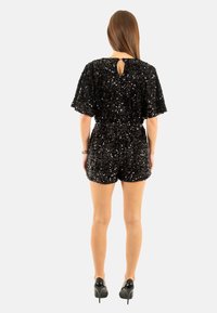 Mono corto de lentejuelas negras con mangas cortas, detalle de abertura en la espalda y cintura elástica. Combinado con stilettos negros, mostrando una textura brillante.