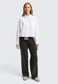 Camicia bianca con bottoni e taschino sul petto, abbinata a pantaloni neri a gamba larga. Un dettaglio a righe si intravede dall'orlo inferiore della camicia. Calzature leggere.