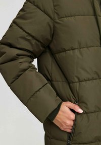 Doudoune olive verte avec des sections matelassées horizontales, poches zippées et une texture douce. Les poignets et les coutures sont cousus pour plus de durabilité.