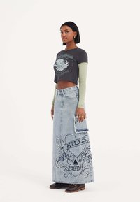 Schwarzes kurzes T-Shirt mit grafischem Druck, über einem langärmligen grünen Oberteil getragen. Blaue Denim-Maxi-Rock mit Schädel- und Herzstickerei, ausgefranster Tasche.
