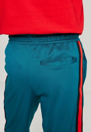 Turkooizen sportbroek met rode en zwarte zijstrepen en een achterzak, gedragen met een rood shirt.