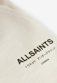 Tessuto beige con una superficie testurizzata, caratterizzato dal logo stampato "ALLSAINTS" e testo in giapponese e "LONDON" in nero.