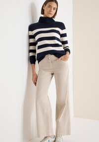 Pull à col roulé rayé bleu marine et cr�ème, associé à un pantalon beige à jambes larges et des baskets claires, avec des boutons décoratifs sur le pantalon.