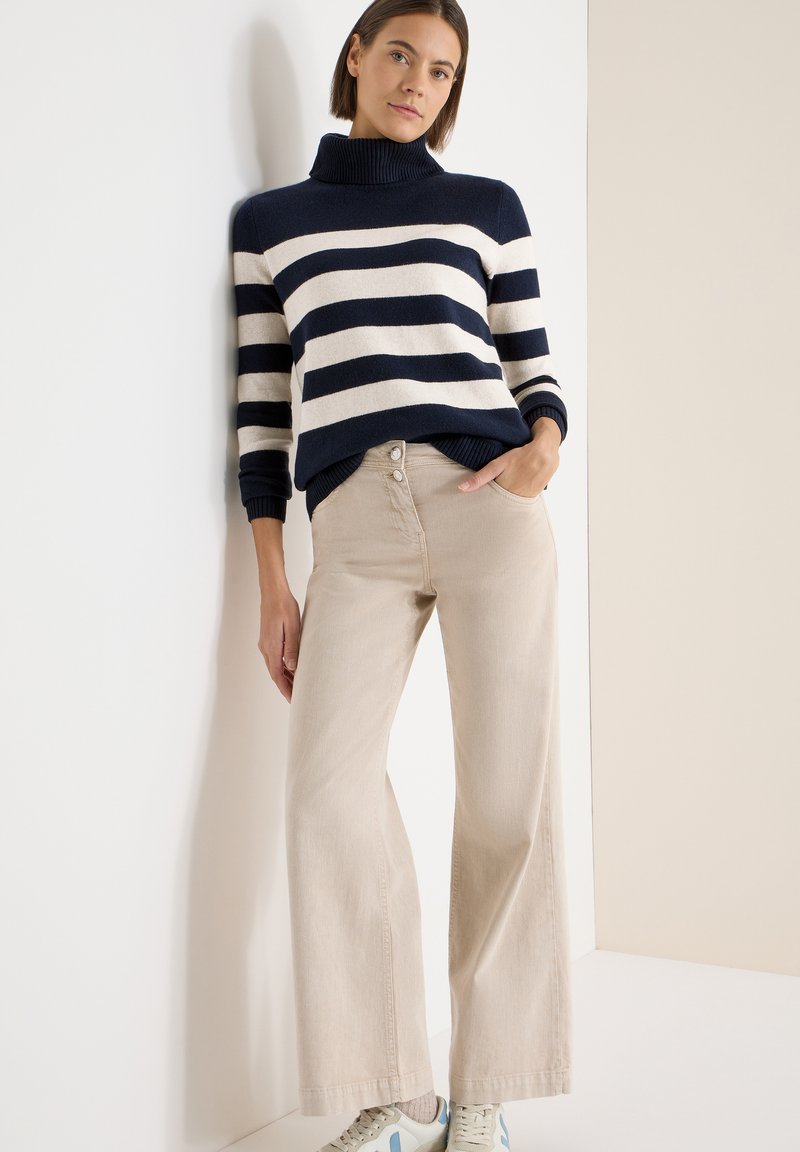 Pull à col roulé rayé bleu marine et crème, associé à un pantalon beige à jambes larges et des baskets claires, avec des boutons décoratifs sur le pantalon.