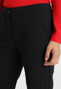 Zwarte op maat gemaakte broeken van een gladde stof, met zijzakken en een slim fit, gecombineerd met een rode top met lange mouwen.