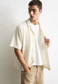 Short-sleeve, light beige shirt with a textured fabric, button front, and a relaxed fit, layered over a white t-shirt and khaki pants.