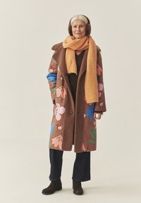 Brązowy płaszcz z kolorowymi kwiatowymi wzorami, zestawiony z pomarańczowym prążkowanym szalikiem. Brązowe botki dopełniają strój. Nauszniki zakrywają uszy.