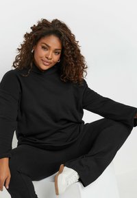 Svart hoodie med hög krage och lös passform, tillsammans med svarta byxor. Modellen bär vita sneakers med en gummisula.