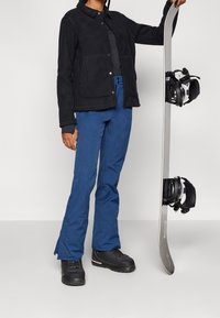 Giacca in pile nera con bottoni a scatto, pantaloni da sci blu e stivali da snowboard neri. Tiene in mano uno snowboard con attacchi neri montati.