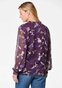 Blusa floreale viola con tessuto trasparente, maniche lunghe e scollatura con balze; presenta motivi bianchi e viola scuro su tutta la superficie.