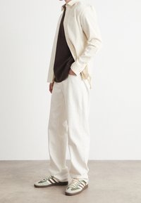 Camicia beige a bottoni sopra una maglietta marrone, abbinata a pantaloni off-white e sneakers con accenti verdi e righe e lacci marroni.