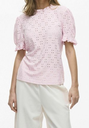 Femme portant un chemisier ajouré rose clair à manches courtes bouffantes et un pantalon blanc à jambes larges sur fond blanc.