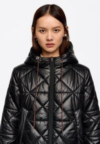 Czarna pikowana kurtka puchowa z kapturem, z ukośnym przeszyciem, sznurkami zakończonymi metalowymi końcówkami oraz zamkiem błyskawicznym z przodu.